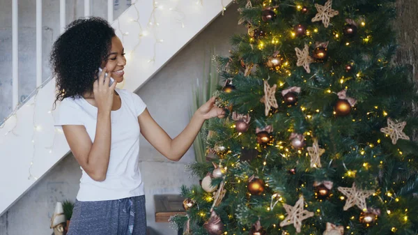 Curly menina de raça mista decorando árvore de Natal em casa e conversando usando smartphone — Fotografia de Stock