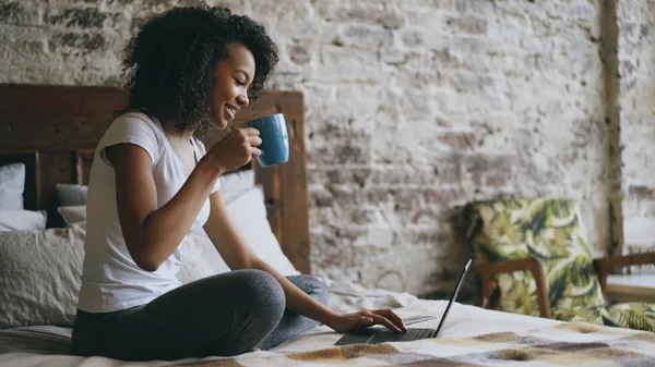 Fröhliches gemischtes Rassenmädchen tippt auf Laptop-Computer, um soziale Medien zu teilen, zu Hause im Bett sitzend — Stockfoto
