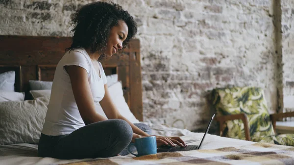 Fröhliches gemischtes Rassenmädchen tippt auf Laptop-Computer, um soziale Medien zu teilen, zu Hause im Bett sitzend — Stockfoto