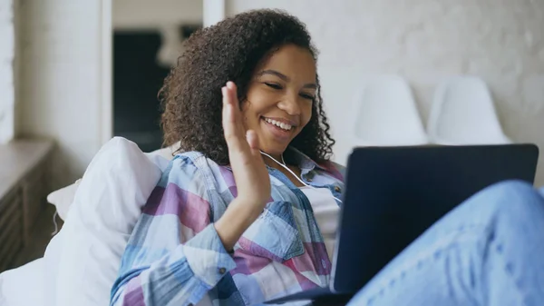 Cheerful mixed race girl having video chat with friends using laptop camera while lying on bed — Stock Photo, Image