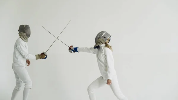 Young woman fencer having fencing training with trainer in white studio indoors