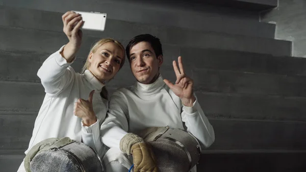 Dos jóvenes esgrimistas sonrientes hombre y mujer tomando selfie en la cámara del teléfono inteligente después del entrenamiento de esgrima en interiores —  Fotos de Stock