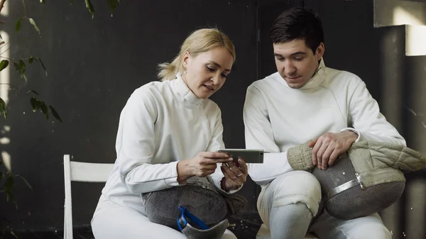 Two young fencers man and woman watching fencing tutorial on smartphone and sharing experience after training indoors — Stock Photo, Image