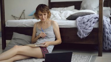 Neşeli genç kadın uygulama ile dizüstü bilgisayar oturma yatak yakınındaki evde kullanarak kalemler kroki çizmek için