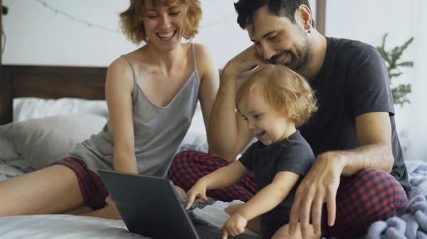 Aranyos kislány, lányom, miközben ő gépelés-ra laptop számítógép ült az ágyban otthon játszó fiatal család — Stock Fotó