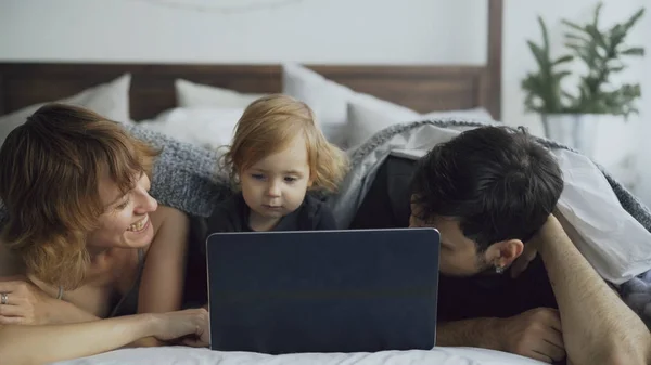 Família jovem com linda menina brincando com a filha enquanto ela assiste computador portátil deitado na cama em casa — Fotografia de Stock