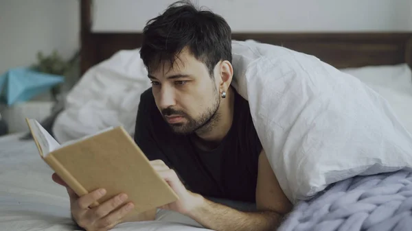 Beau jeune homme lisant un livre couché au lit sous la couverture à la maison — Photo