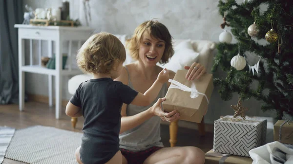 Feliz madre y su pequeña hija desempaquetan la caja de regalo cerca del árbol de Navidad en casa — Foto de Stock