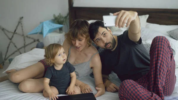 Молодая семья с симпатичной маленькой девочкой, делающей селфи на камеру смартфона сидя дома в постели — стоковое фото
