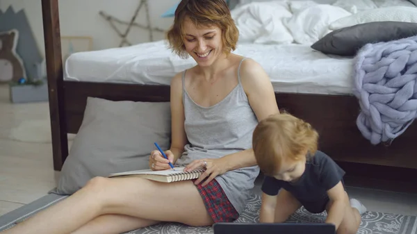 Mère heureuse avec mignon dessin adorable fille dans l'album avec des crayons assis près du lit à la maison — Photo