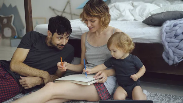 Счастливая семья с симпатичной дочкой, рисующей в альбоме с ручкой, сидящей у кровати дома — стоковое фото