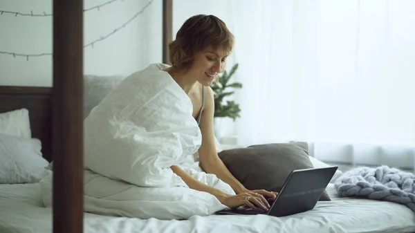 Jovem alegre usando seu computador portátil sentado na cama em casa — Fotografia de Stock
