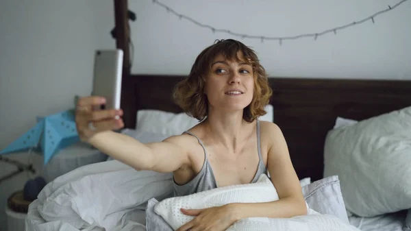 Fiatal vidám nő vesz használ smartphone ült az ágyban, az otthon selfie-portré — Stock Fotó