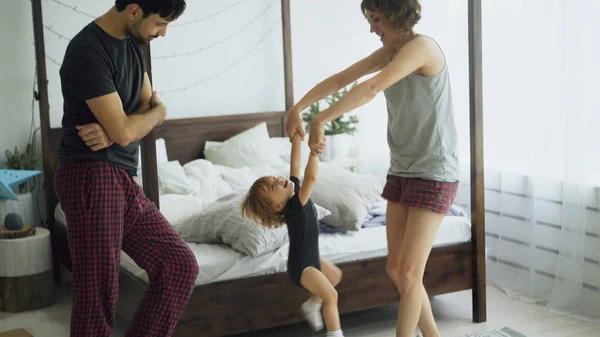Familia feliz y pequeña hija linda bailando y girando cerca de la cama en el dormitorio en casa — Foto de Stock