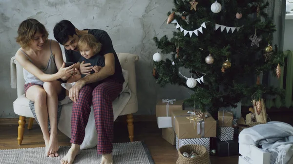 Glückliche Familie mit süßer kleiner Tochter sitzt am Weihnachtsbaum und nutzt Smartphone zu Hause — Stockfoto