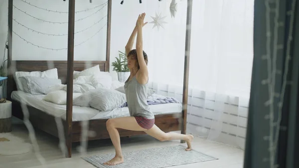 Giovane bella donna che fa esercizio di yoga vicino a letto in camera da letto a casa — Foto Stock