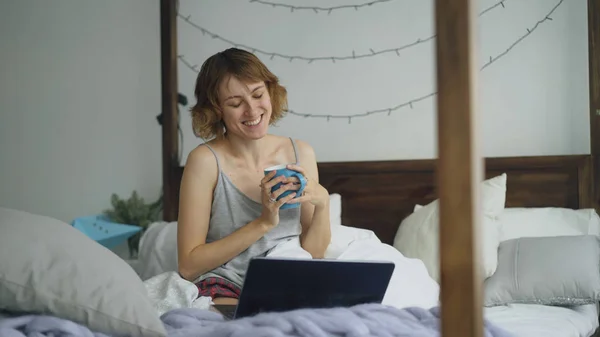 Mulher alegre atraente ter bate-papo de vídeo on-line com amigos usando câmera laptop enquanto sentado na cama em casa — Fotografia de Stock