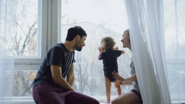 Familia feliz con joven linda hija sentada en el alféizar de la ventana jugando y mirando en la ventana en casa — Foto de Stock