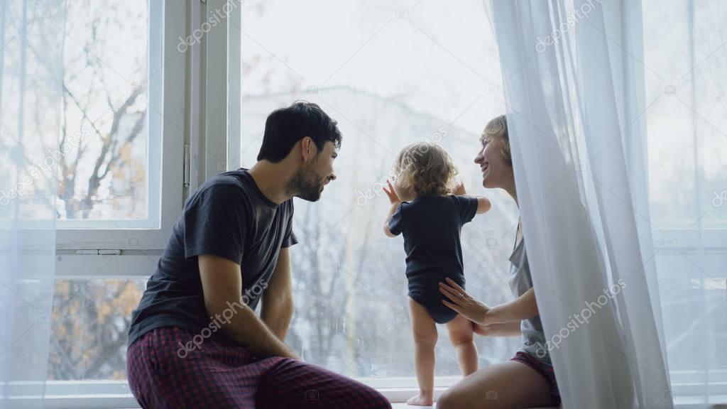 Happy family with young cute daughter sitting on windowsill playing and looking in window at home