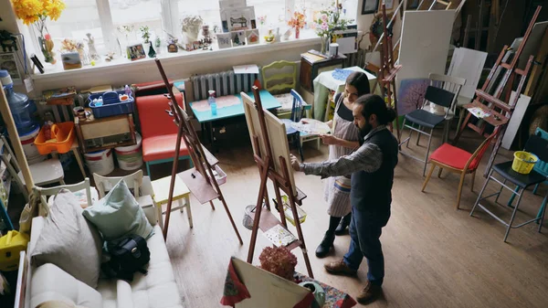 Artista qualificado homem ensinando jovem mulher pintura no cavalete no estúdio da escola de arte - criatividade, educação e arte pessoas conceito — Fotografia de Stock