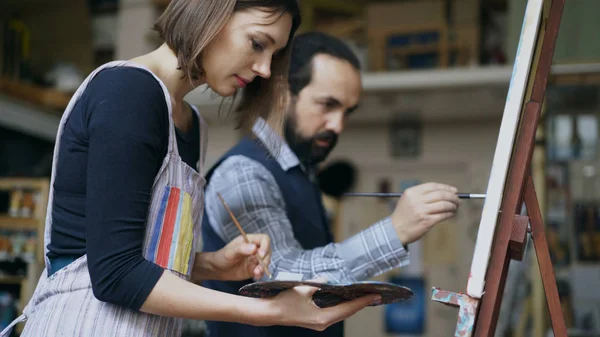 Artiste compétent homme enseignant la peinture jeune femme sur chevalet à l'atelier de l'école d'art - concept de créativité, d'éducation et de gens de l'art — Photo