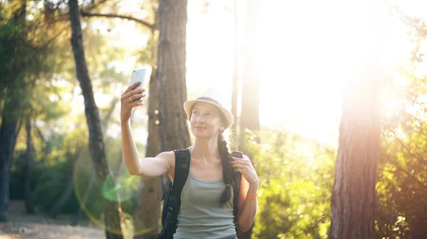 Забавная туристка в шляпе делает селфи с камерой на смартфоне во время путешествия и автостопом — стоковое фото