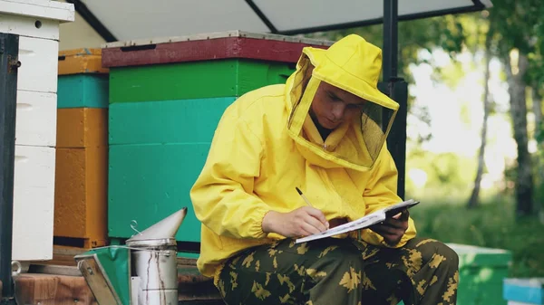 Pszczelarz młody człowiek napisać w Notatniku sprawdzanie zbiorów siedząc w pobliżu ule — Zdjęcie stockowe