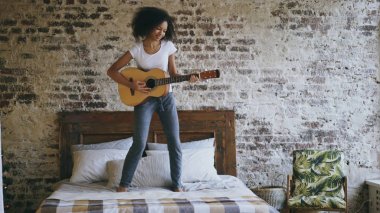 Karışık yarış genç komik kız akustik gitar çalmak ve yatağa evde dans eğlenceli