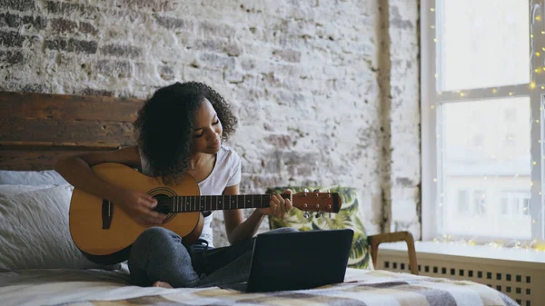 Rizado afroamericano adolescente chica concéntrica aprender a tocar la guitarra usando ordenador portátil sentado en la cama en casa — Foto de Stock