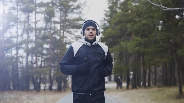 Attraente corridore uomo in cuffia che fa jogging mentre ascolta musica nel parco invernale al mattino — Foto Stock