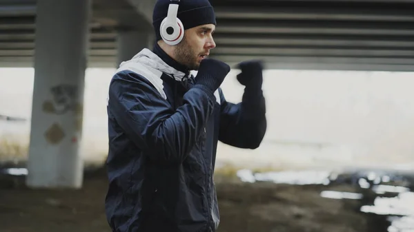 Boxeador deportivo en auriculares haciendo ejercicio de boxeo en ubicación urbana al aire libre en invierno — Foto de Stock