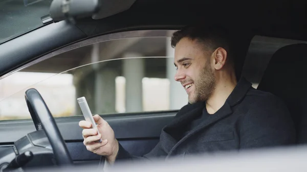 Привлекательный бизнесмен сидит в машине и смеется, пользуясь смартфоном после поездки — стоковое фото