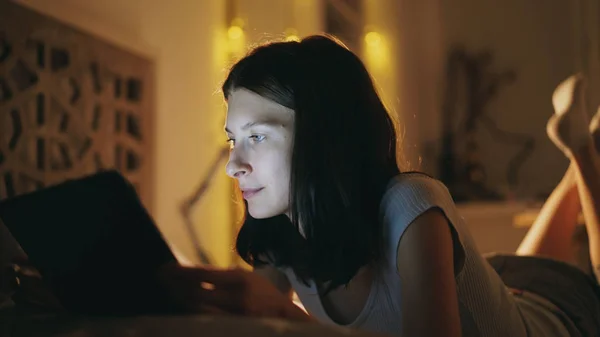 Junge lächelnde Frau nutzt Tablet-Computer, um soziale Medien zu teilen, wenn sie nachts zu Hause im Bett liegt — Stockfoto