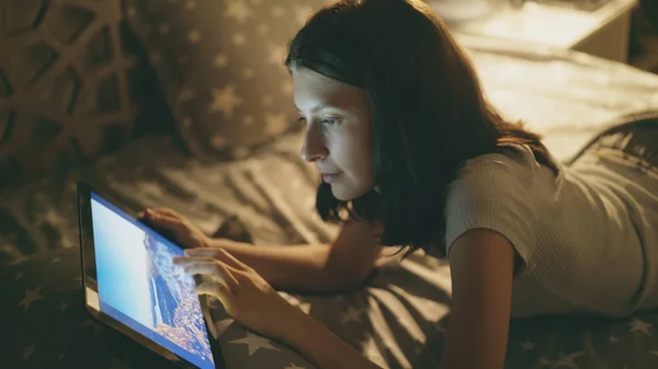 Молодая улыбающаяся женщина лежит на кровати и смотрит фотографии из путешествия на цифровом планшетном компьютере ночью дома — стоковое фото