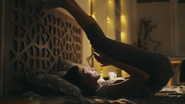 Mulher sorridente feliz usando smartphone para surfar nas redes sociais enquanto faz exercício de ioga deitado na cama à noite — Fotografia de Stock