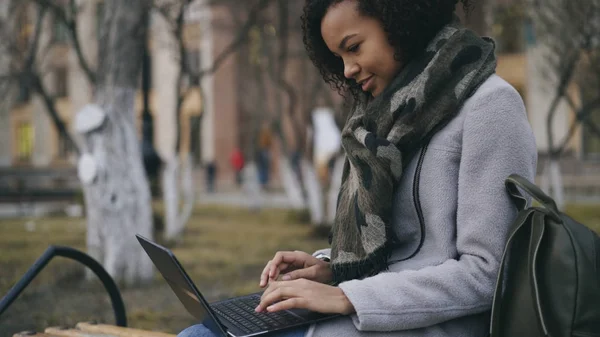 Attraktive afrikanisch-amerikanische Studentin tippt auf Laptop-Computer sitzt auf Bank in der Nähe von Univercity — Stockfoto