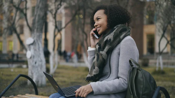 Attraktive afrikanisch-amerikanische Studentin telefoniert auf Bank sitzend mit Laptop-Computer im Freien — Stockfoto