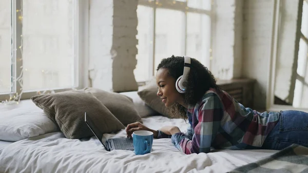 Atraktivní smíšený závod dívka poslouchat hudbu při procházení sociální média na notebooku ležící na posteli — Stock fotografie
