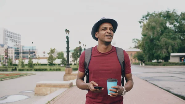 Steadicam снимок улыбающегося студента в шляпе, гуляющего и серфингирующего смартфон, пьющего кофе на улице — стоковое фото