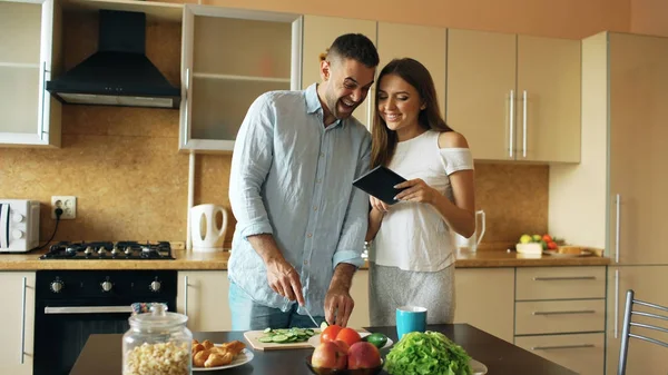 Atractiva pareja se reúnen en la cocina temprano en la mañana. Mujer guapa usando tableta compartiendo su marido en las redes sociales — Foto de Stock