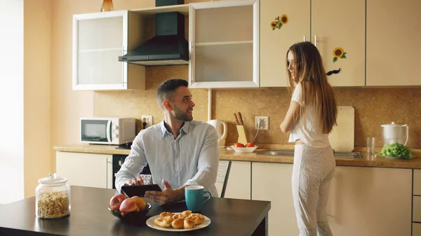 Jovem feliz usando computador tablet digital sentado na cozinha e falando com sua esposa enquanto ela cozinha em casa — Fotografia de Stock