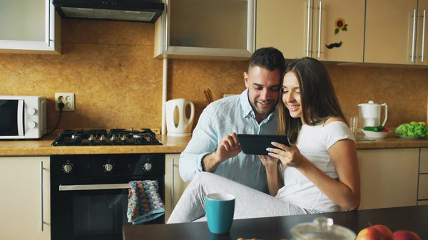 Glückliches junges Paar mit digitalem Tablet-Computer, während es morgens in der Küche sitzt und frühstückt — Stockfoto