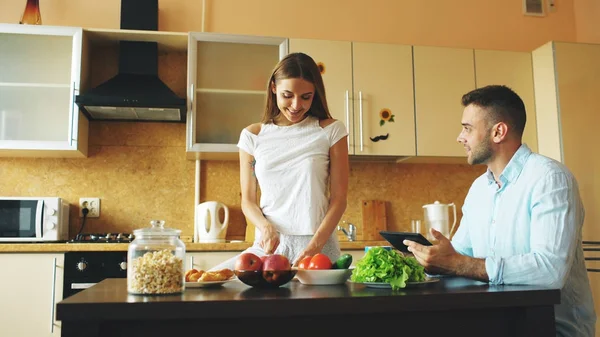 Attraktives Paar plaudert frühmorgens in der Küche. schöner Mann mit Tablette, während seine Freundin kocht — Stockfoto