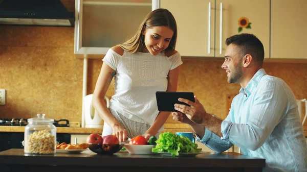 Привлекательная пара болтает на кухне рано утром. Красивый человек, использующий планшет, пока его девушка готовит — стоковое фото