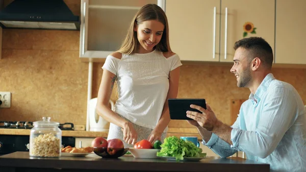 Привлекательная пара болтает на кухне рано утром. Красивый человек, использующий планшет, пока его девушка готовит — стоковое фото