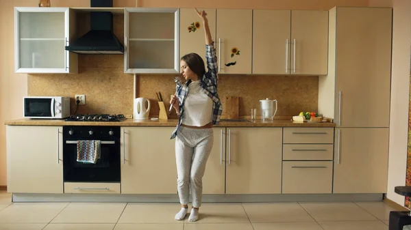 Vidám fiatal vicces nő táncol és énekel, kanál, miközben szabadidő otthon a konyhában — Stock Fotó