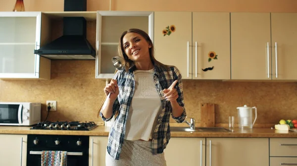 Веселая юная забавная женщина танцует и поет с ковшами во время досуга на кухне дома — стоковое фото