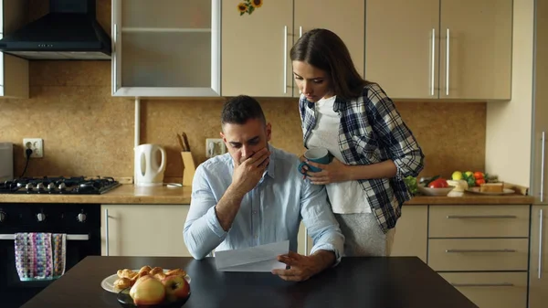 Молодий чоловік читає неоплачені рахунки і обіймається дружиною, яка підтримує його на кухні вдома — стокове фото