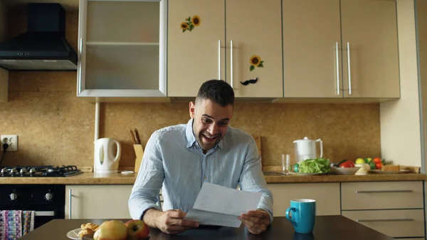 Красавчик получает хорошие новости, читая письмо на кухне, пока завтракает дома рано утром — стоковое фото