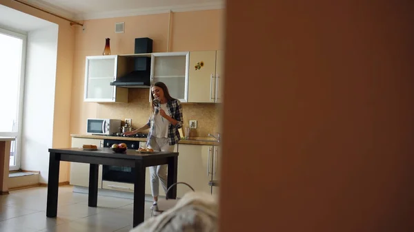 Vrolijke jonge grappige vrouw dansen en zingen met pollepel terwijl tijdens het koken in de keuken thuis — Stockfoto